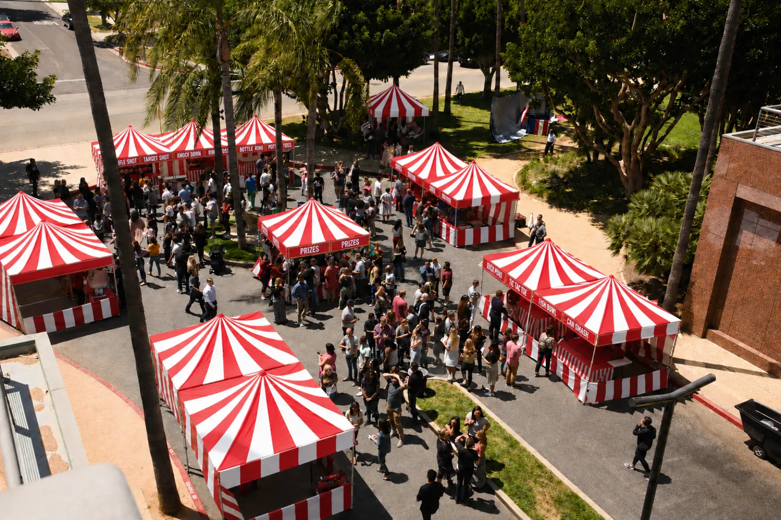 A wide overhead view of a public event midway, striped carnival booths and prize stands framing a paved plaza busy with guests in Lancaster, CA