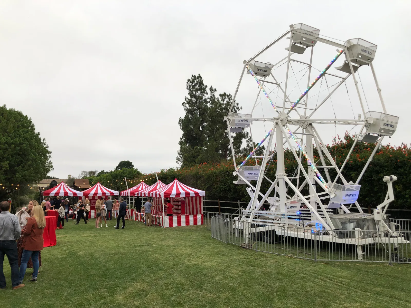 A white Ferris wheel strung with lights on a lawn beside a row of red-and-white striped game booths at a neighborhood event
