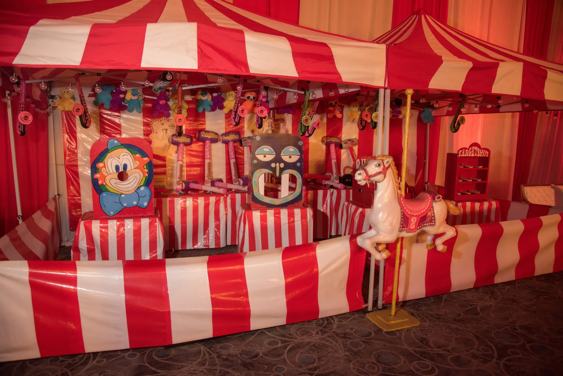 A wider view of multiple striped 10x10 carnival booths in a midway layout on a parking lot, with prize plush hanging visibly from each booth's internal frame