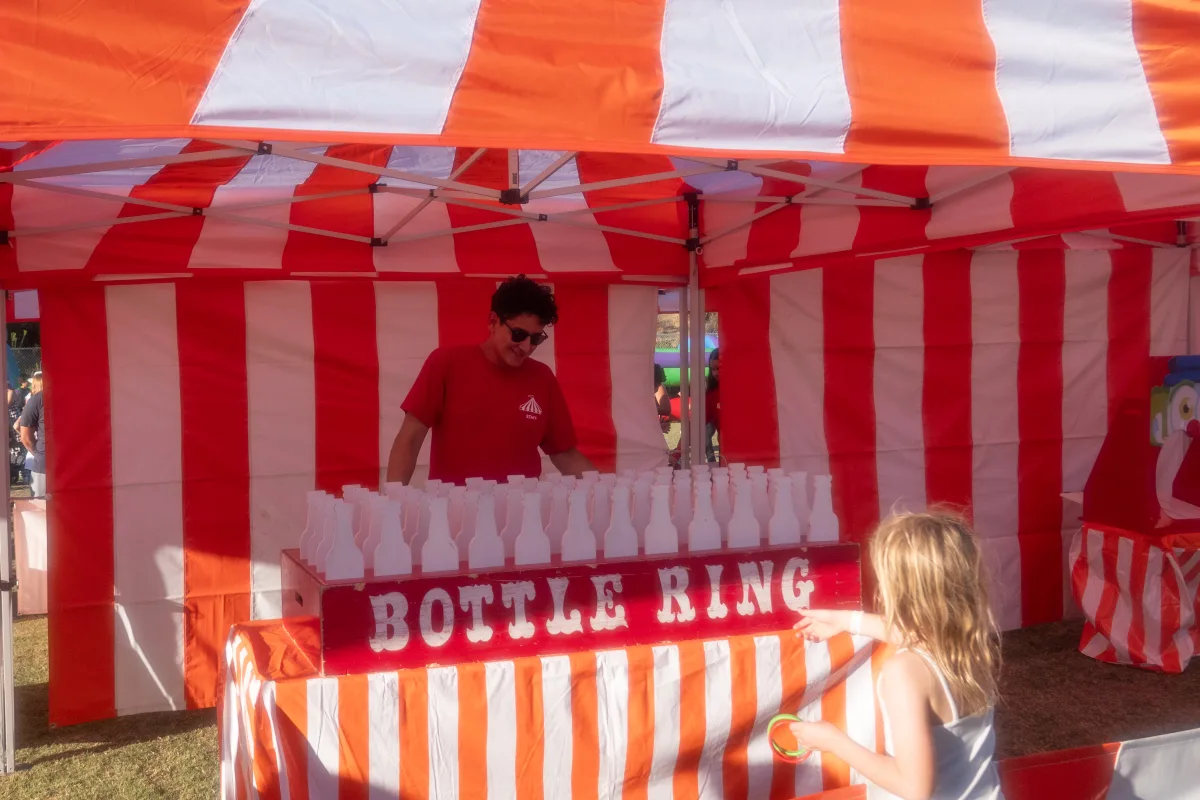 A child playing a Bottle Ring game at an orange-and-white striped carnival booth, one of the Halloween booth colors offered for La Cañada Flintridge events
