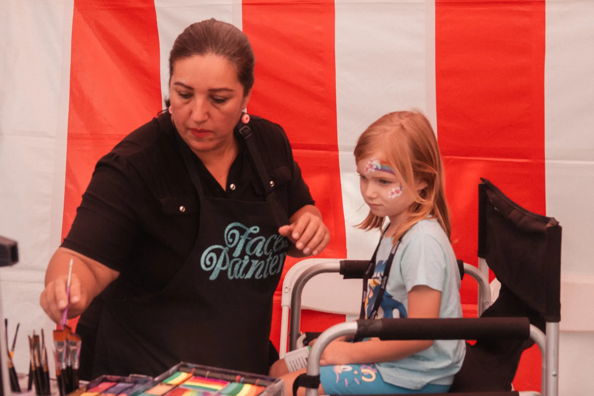 A row of carnival birthday games with plush prizes, a ring toss booth, and a concession table set up on a paved outdoor area