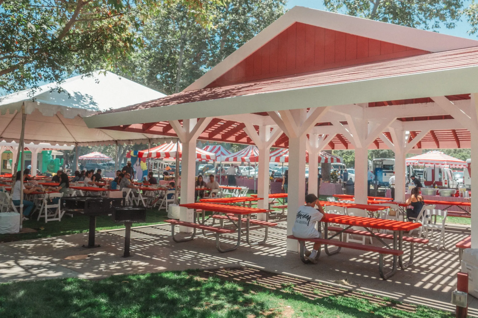 A corporate family day spread across a large open lot with multiple striped carnival game booths, a bounce house obstacle course in the background, and employees and children gathered near concession stations in Carson, CA