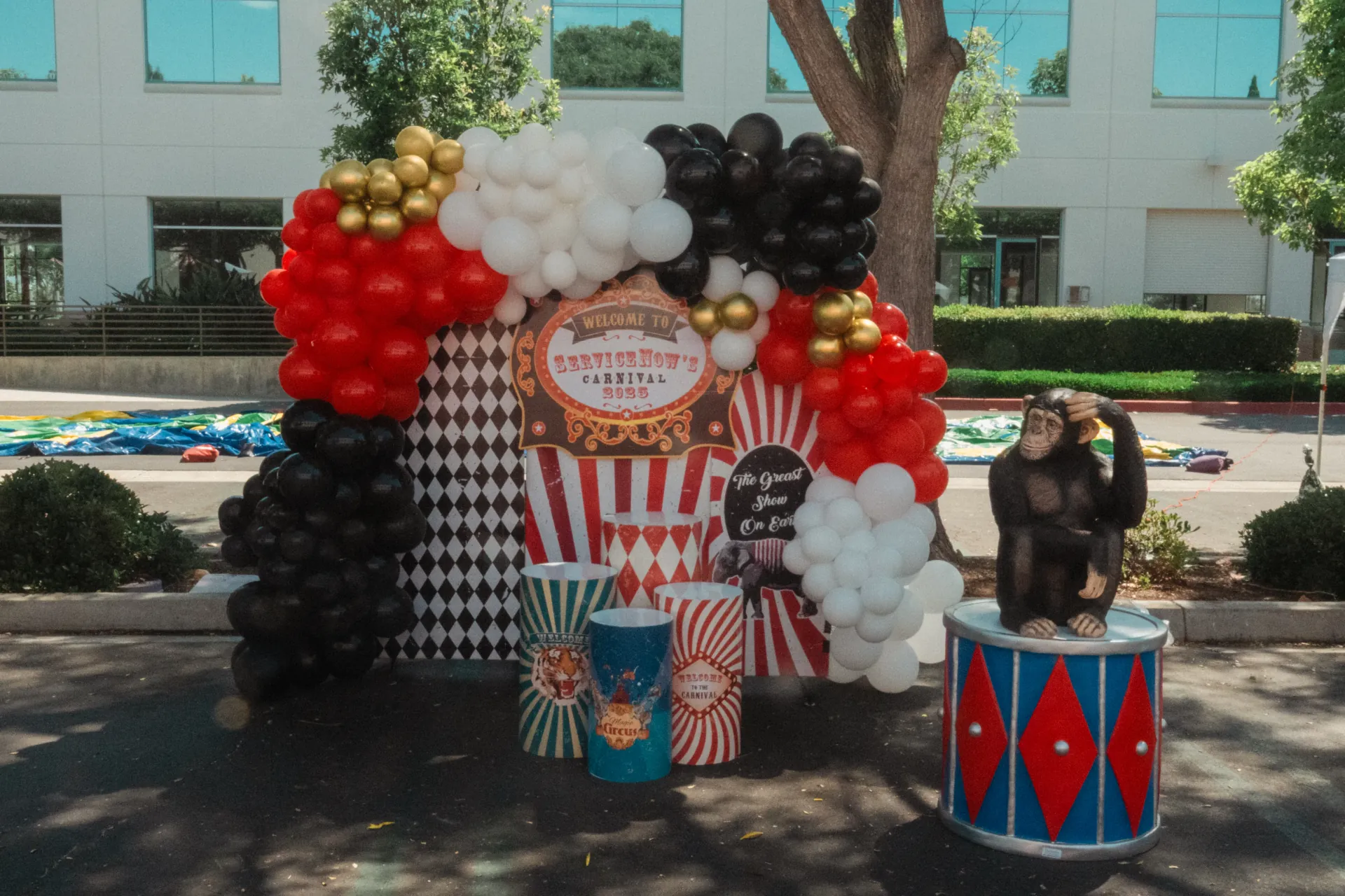 A row of staffed carnival game booths at a large outdoor corporate event with employees and children queued at each station and prize plush hanging above the booth openings