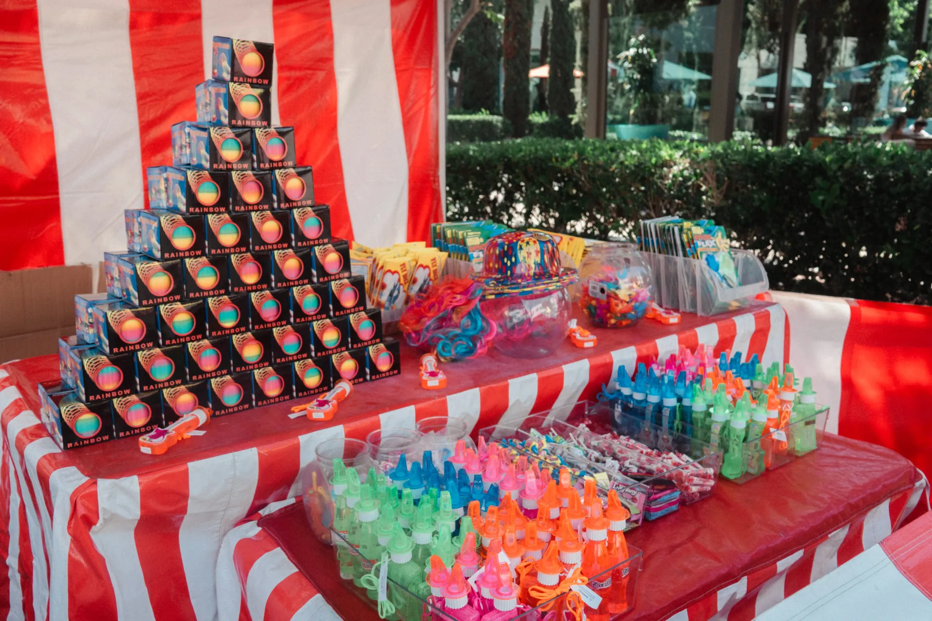 An attendant in a striped vest running a carnival game booth at a community fundraiser, with guests lining up and prize plush hanging above the counter