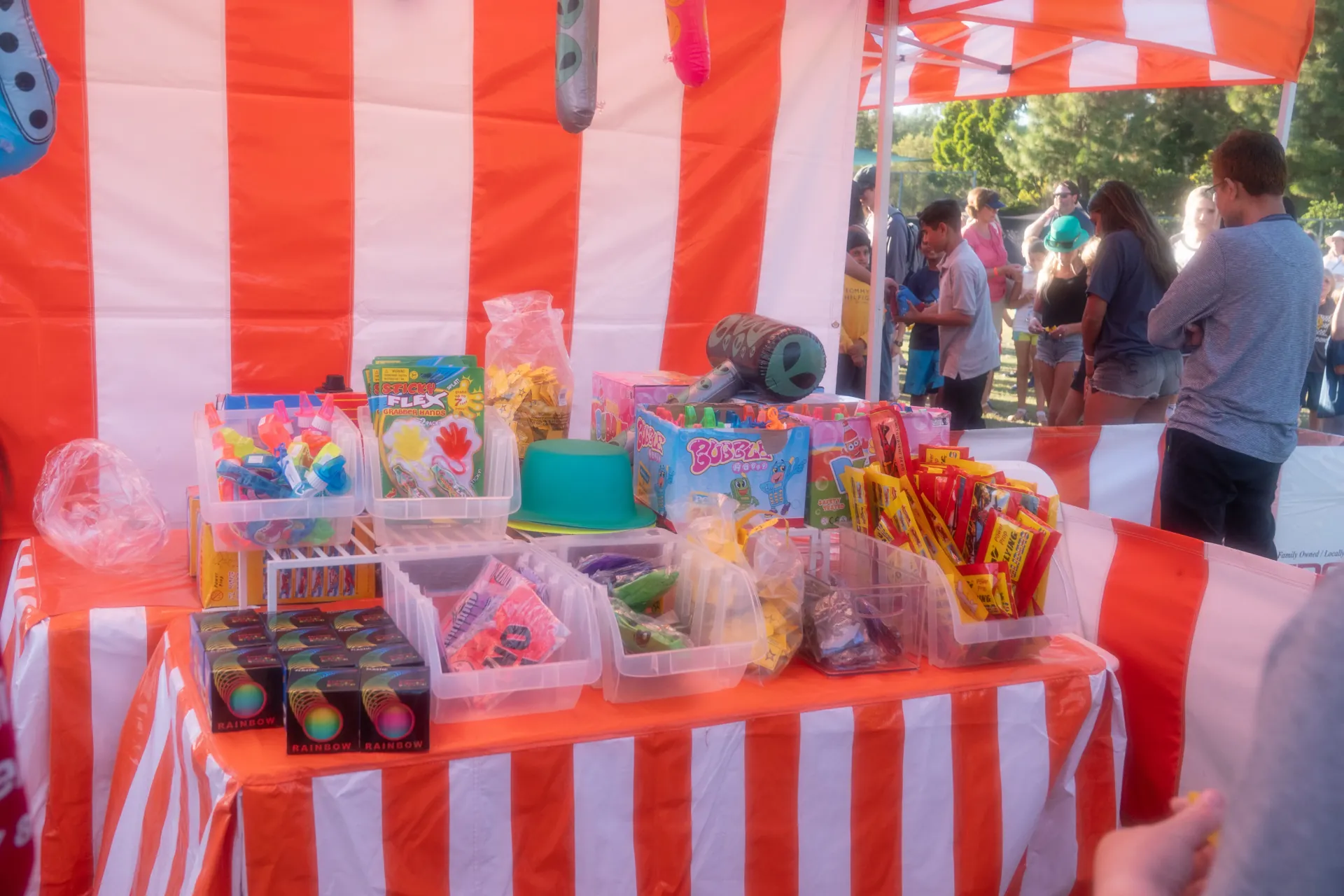 A row of carnival game booths at a park fundraiser, with striped tents, prize displays visible above the counters, and families playing under an open sky