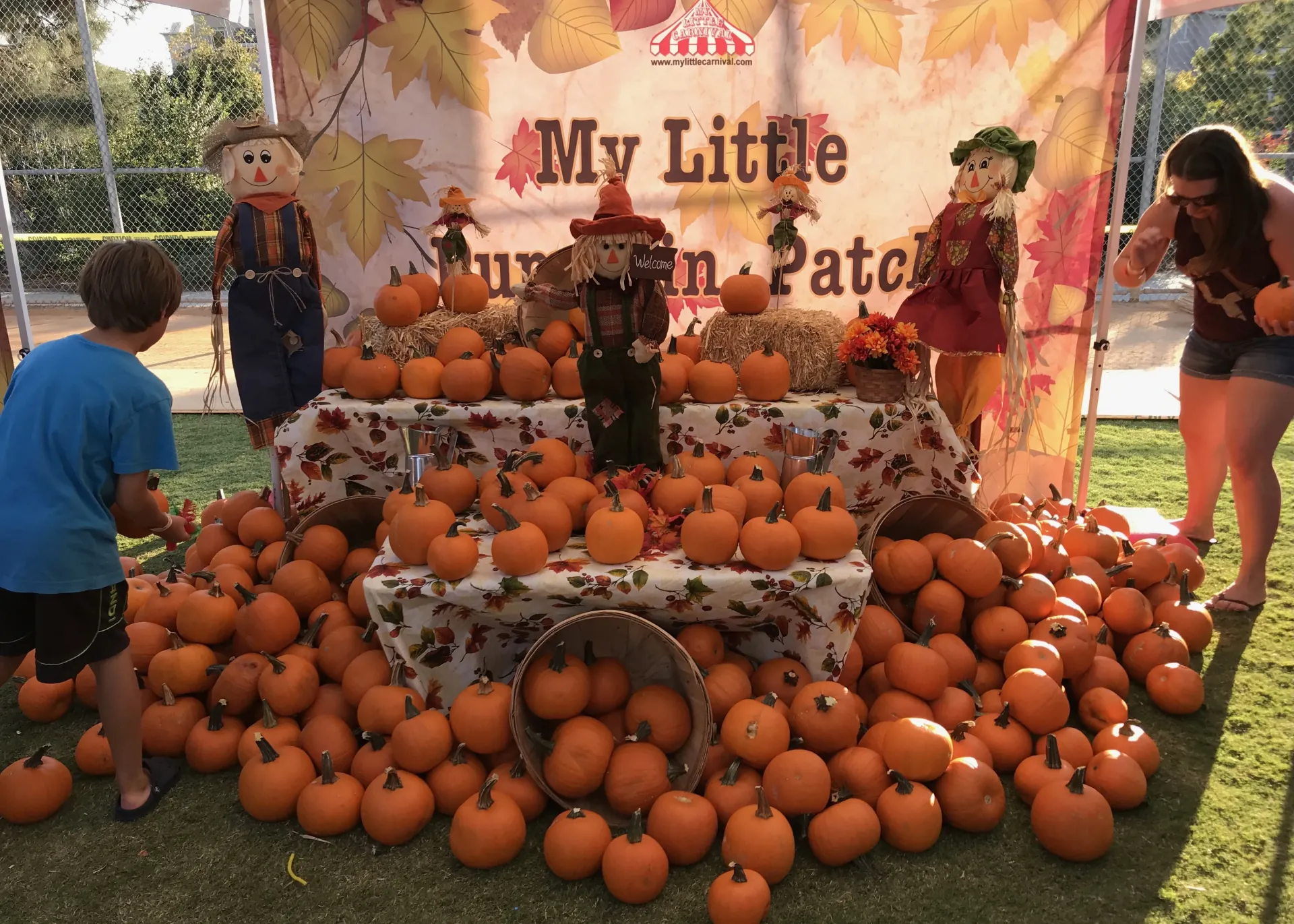 A holiday-themed carnival booth with garland and ornament decorations, an attendant in a festive vest handing a prize to a child in a winter hat