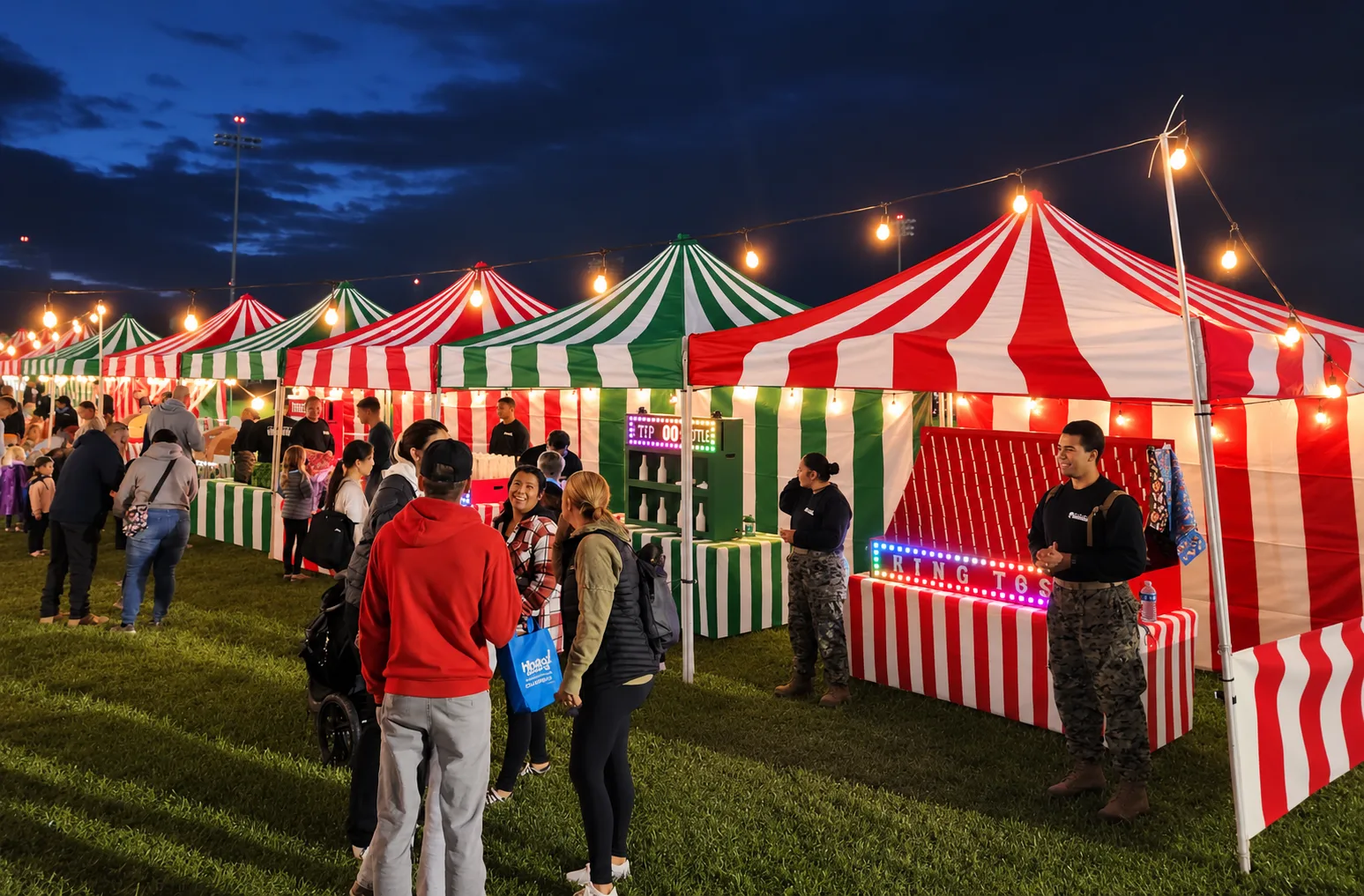 Red-and-white and green-and-white striped carnival booths under string lights at dusk, with a lit ring-toss marquee, at an outdoor winter holiday event in Carson, CA