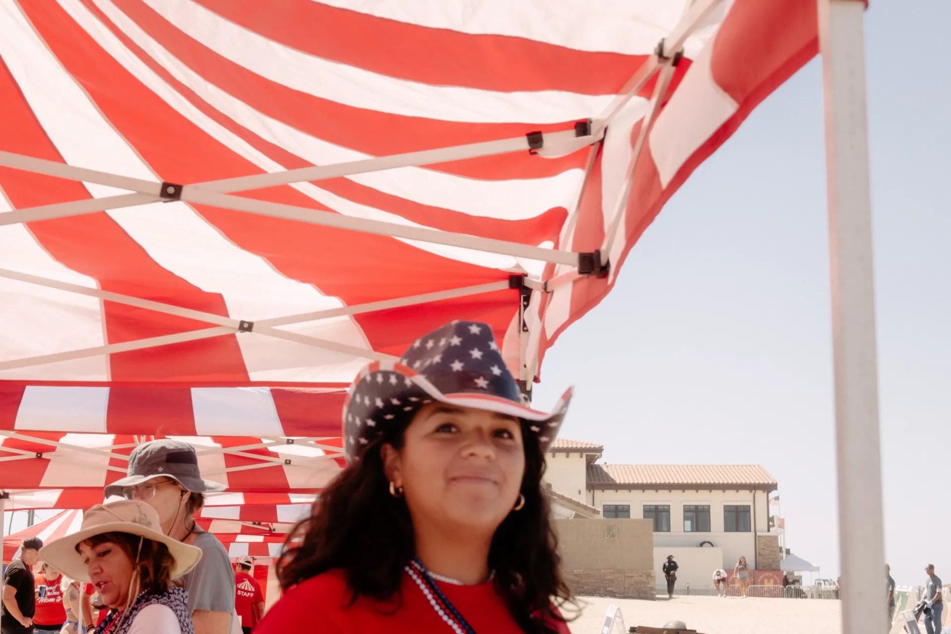 A large outdoor military family day with striped carnival game booths, an inflatable obstacle course, and concession stations spread across a wide grass field with service members and families gathered throughout in Carson, CA