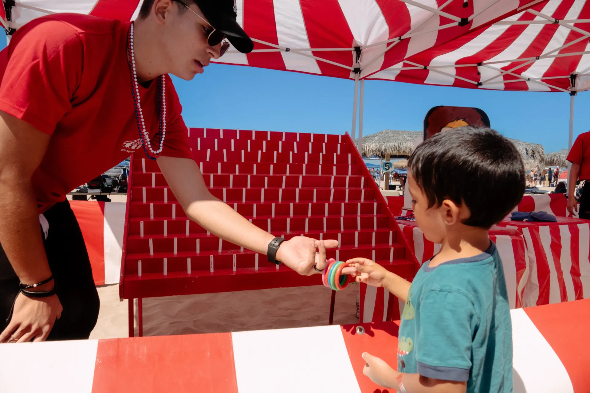 Striped carnival game booths and a popcorn concession station set up on a grass field at a military community appreciation event, with service members and children playing games in the foreground
