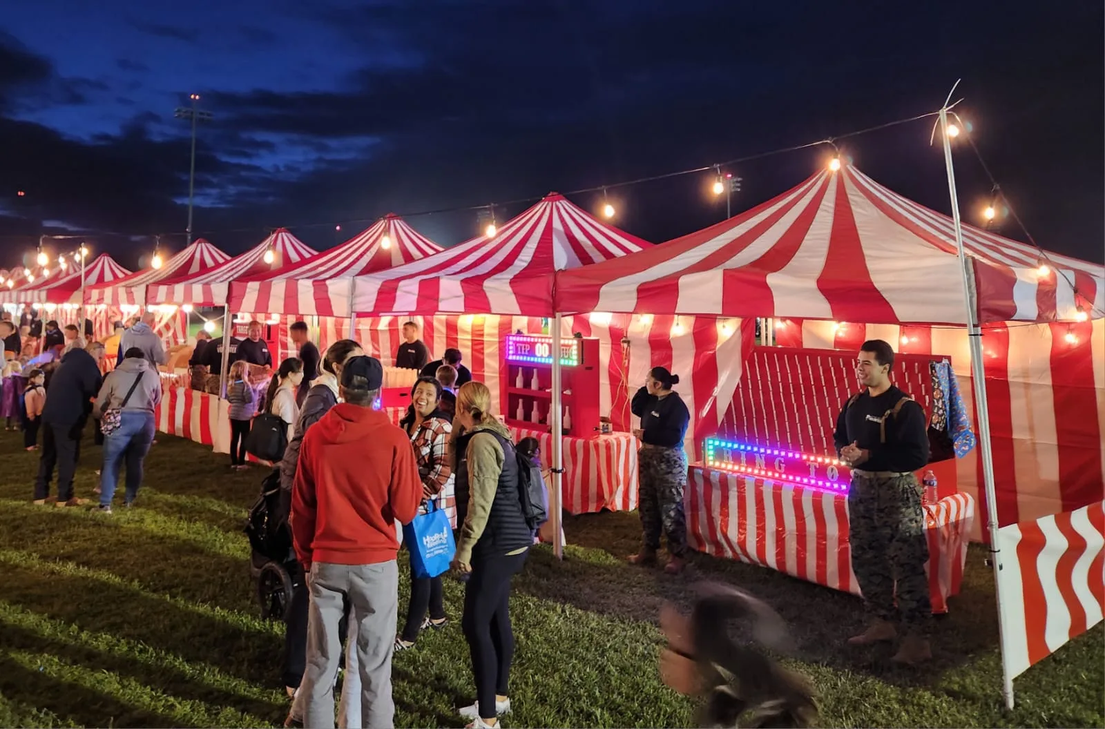 An elementary school carnival on a blacktop with multiple red-and-white striped booths, families lined up, and balloon arches at the entrance in Carson, CA